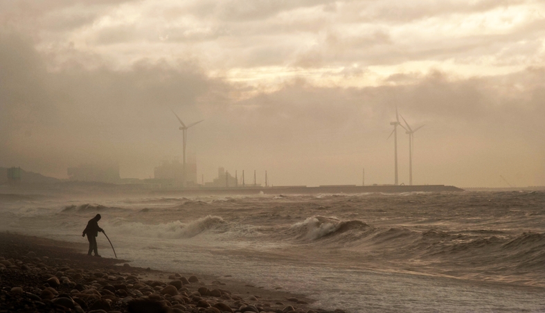 A beach, clouded with air pollution. In the background, windmills are barely visible through the haze.